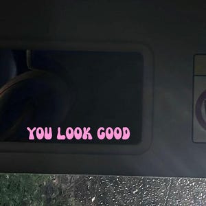 Peut inclure: Un autocollant rose avec les mots "YOU LOOK GOOD" dans une police rétro est collé sur un pare-soleil de voiture. Le texte est clair et centré. L'arrière-plan est sombre, avec une vue sur une fenêtre pluvieuse.