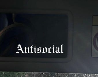 Pode incluir: Um decalque branco "Antisocial" em fonte gótica é exibido em uma superfície escura, possivelmente uma viseira de carro. A imagem é tirada de dentro de um veículo, com gotas de chuva visíveis na janela.