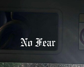 Pode incluir: Um decalque branco com as palavras "No Fear" em uma fonte gótica é exibido em uma viseira de carro. A imagem é tirada de dentro do carro, com uma vista do exterior através do para-brisa.