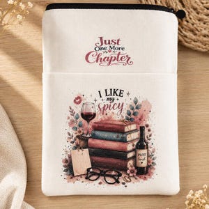 Peut inclure: Une pochette zippée en toile blanche avec le texte Just One More Chapter en haut. En dessous, un graphique montre une pile de livres, un verre de vin rouge, une bouteille étiquetée Plot Twist et des lunettes noires, avec le texte I LIKE my spicy.