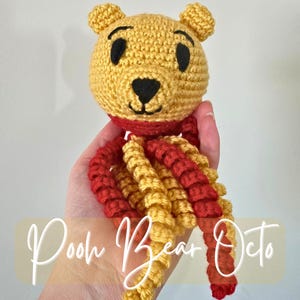 May include: A handmade crochet toy featuring a yellow bear head with black eyes and nose, wearing a red scarf. The bear's body transitions into red and yellow tentacles. The text "Pooh Bear Octo" is written in a cursive font.