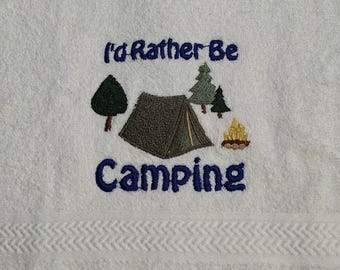 May include: White towel with embroidered camping scene. The design includes a tent, trees, and a campfire, with the text "I'd Rather Be Camping" in blue thread. The towel has a textured border.