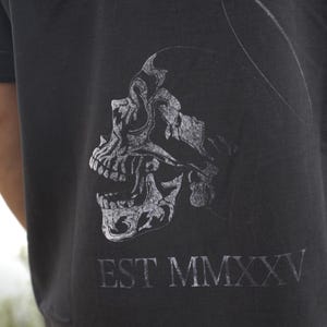 Może przedstawiać: Ciemnoszara koszulka z detaliczną, stylizowaną grafiką czaszki w jaśniejszym odcieniu szarości. Tekst "EST MMXXV" jest wydrukowany pod czaszką. Koszulka ma kr&oacute;tkie rękawy.