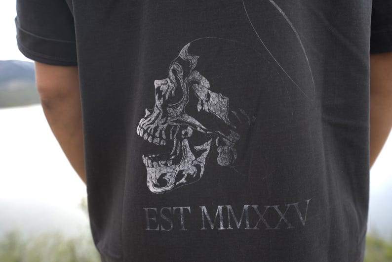 Może przedstawiać: Ciemnoszara koszulka z detaliczną, stylizowaną grafiką czaszki w jaśniejszym odcieniu szarości. Tekst "EST MMXXV" jest wydrukowany pod czaszką. Koszulka ma kr&oacute;tkie rękawy.