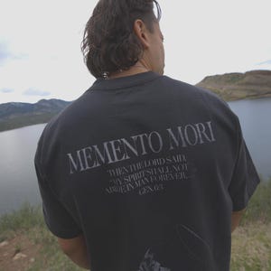 Może przedstawiać: Ciemnoszara koszulka z napisem "MEMENTO MORI" z tyłu w jasnoszarym kolorze. Poniżej znajduje się cytat z Księgi Rodzaju. Koszulka jest noszona przez osobę stojącą nad jeziorem.