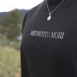 Może przedstawiać: Czarna bluza z okrągłym dekoltem z białym napisem &bdquo;MEMENTO MORI&rdquo;. Widoczny srebrny naszyjnik z okrągłą zawieszką. Tło przedstawia rozmyty krajobraz z drzewami i wodą.