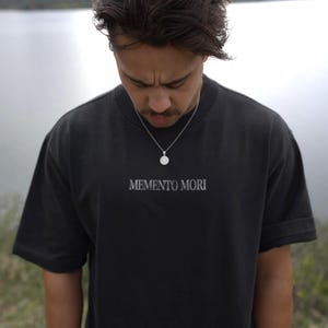 Może przedstawiać: Czarny t-shirt z napisem "MEMENTO MORI" w białym kolorze. Koszulka ma kr&oacute;tkie rękawy i okrągły dekolt. Noszony jest srebrny naszyjnik z wisiorkiem. Tło to rozmyty widok na jezioro.