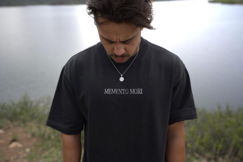 Może przedstawiać: Czarny t-shirt z napisem "MEMENTO MORI" w białym kolorze. Koszulka ma kr&oacute;tkie rękawy i okrągły dekolt. Noszony jest srebrny naszyjnik z wisiorkiem. Tło to rozmyty widok na jezioro.