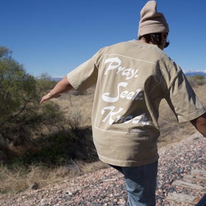 May include: A tan short-sleeved t-shirt with the words "Pray. Seek. Know." in white script. The person is wearing a tan beanie and blue jeans. The background includes a blue sky, trees, and a mountain range.