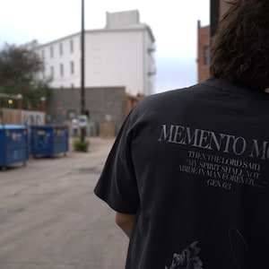 Może przedstawiać: Ciemnoszara koszulka z napisem "MEMENTO MORI" wydrukowanym na plecach stylizowaną czcionką. Poniżej tekstu znajduje się cytat z Księgi Rodzaju. Koszulka jest noszona przez osobę opartą o ceglany mur.