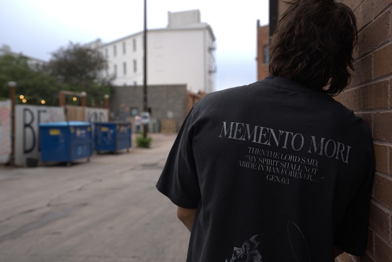 Może przedstawiać: Ciemnoszara koszulka z napisem "MEMENTO MORI" wydrukowanym na plecach stylizowaną czcionką. Poniżej tekstu znajduje się cytat z Księgi Rodzaju. Koszulka jest noszona przez osobę opartą o ceglany mur.