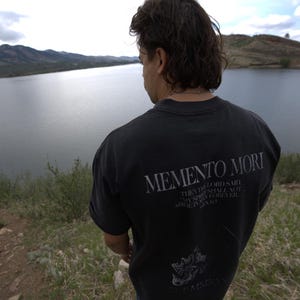 Może przedstawiać: Czarny t-shirt z napisem "MEMENTO MORI" z tyłu. Poniżej tekstu znajduje się cytat i grafika. Osoba stoi na trawiastym wzg&oacute;rzu z widokiem na jezioro i g&oacute;ry.