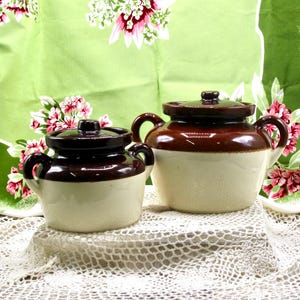May include: Two ceramic pots with lids, each featuring a two-tone design with a dark brown glazed upper section and a cream-colored lower body. Both pots have two side handles and a small knob on the lid, showcasing a classic stoneware aesthetic.