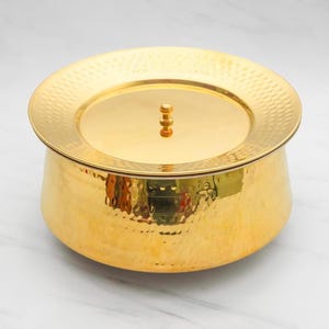 May include: A gold-colored serving bowl with a matching lid. The bowl has a hammered texture and a rounded shape. The lid features a small, decorative handle. The bowl is likely made of metal and is suitable for serving food.
