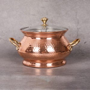 May include: A round, hammered copper pot with a wide base and a tapered neck. It features two curved brass side handles and a clear glass lid topped with a small, polished brass knob. The exterior surface has a textured, dimpled finish.