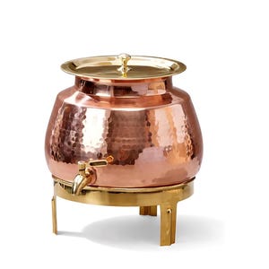 May include: A hammered copper beverage dispenser with a polished gold-tone lid and base. The round vessel features a matching gold-tone spigot on the front, a wide opening at the top, and a sturdy stand with three legs for elevated placement.