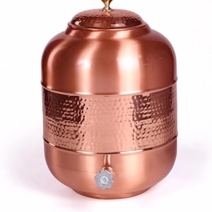 Puede incluir: Un dispensador de agua de cobre con una banda de textura martillada. El dispensador tiene una tapa con un pomo dorado y un grifo plateado. El acabado de cobre le da al dispensador un aspecto cálido y acogedor.