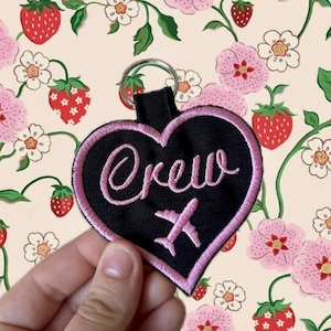 May include: A heart-shaped black fabric keychain with a pink embroidered border. The word "crew" is embroidered in pink cursive script above a small pink embroidered airplane icon. A silver metal split ring is attached to the top of the heart.