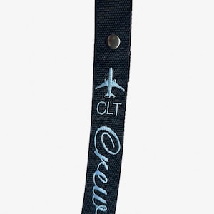May include: A black fabric luggage tag featuring a silver metal snap button at the top. The tag is embroidered with a light blue airplane icon, the letters CLT, and the word Crew in a cursive script font oriented vertically along the strap.
