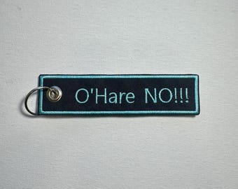 May include: Black rectangular keychain with the text "O'Hare NO!!!" embroidered in teal. The keychain has a teal border and a silver metal ring attached to the left side. The background is a white textured surface.