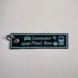 May include: Black rectangular keychain with teal embroidered text and icons. The text reads "Commuter Final Boss" with icons of a car, airplane, train, bus, and ship. A silver key ring is attached to the top left corner.