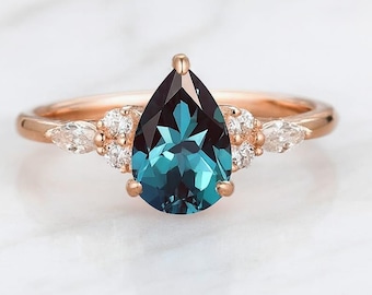 May include: Rose gold ring featuring a pear-shaped, teal-colored gemstone. The ring is accented with small, clear stones on either side of the main stone. The band is a delicate rose gold color, creating an elegant and sophisticated look.
