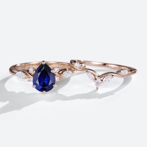 May include: Two rose gold rings. One ring features a pear-shaped, deep blue gemstone surrounded by smaller white gemstones. The second ring has a curved design with multiple white gemstones.