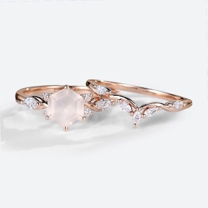 May include: A rose gold ring set. The main ring has a hexagonal rose quartz centre stone and small white gemstones. The second ring has a curved design with leaf-like accents and white gemstones. The set is delicate and elegant.
