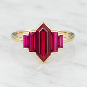 May include: A gold ring featuring a central, vibrant red gemstone with a geometric, Art Deco design. The ring's band is a polished gold, and the gemstone is flanked by smaller, matching red stones.
