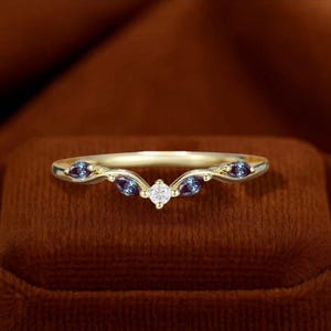 May include: A gold ring with a curved design. The ring features three dark blue-purple gemstones and a small, round, clear gemstone in the centre. The ring is set on a brown surface.