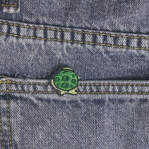 Puede incluir: Primer plano de un bolsillo de mezclilla con un pequeño parche de tortuga verde. El parche está bordado con un diseño detallado de caparazón y un contorno negro. La mezclilla es azul con costuras amarillas.