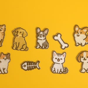 May include: Embroidered patches featuring various cat and dog designs, including a tabby cat, a golden retriever, a French bulldog, a corgi, and a beagle. Also includes a bone and fishbone patch. All patches are on a yellow background.
