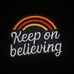 Puede incluir: Parche bordado con un arcoíris naranja, rojo y amarillo, con las palabras "Keep on believing" en escritura blanca. Sobre un fondo negro.