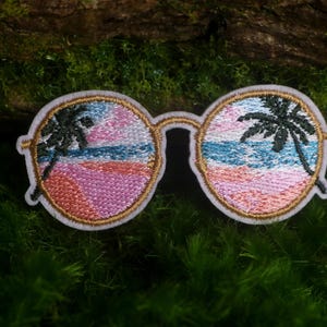 May include: An embroidered patch shaped like round sunglasses with gold-colored frames. The lenses feature a detailed stitched scene of a tropical beach with a blue ocean, a pink sandy shore, and a dark green palm tree silhouette in each lens.