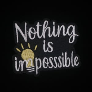 Puede incluir: Parche bordado con las palabras "Nothing is impossible" en escritura blanca. Una bombilla amarilla con rayos está debajo de las palabras. El parche está sobre un fondo negro.