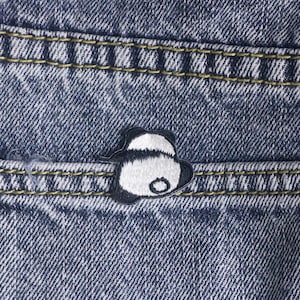 Könnte beinhalten: Nahaufnahme einer Jeans-Tasche mit einem kleinen Panda-Aufnäher. Der Aufnäher ist schwarz-weiß und die Tasche hat gelbe Nähte. Der Denim hat eine verwaschene blaue Farbe.