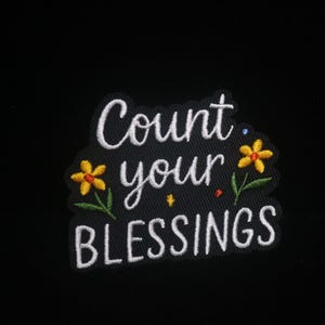 Op de afbeelding: Zwart geborduurde patch met de witte tekst "Count your Blessings". Voorzien van gele en groene bloemaccenten. Perfect voor het toevoegen van een positieve boodschap aan kleding of accessoires.