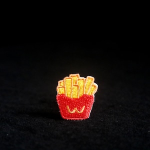 以下が含まれることがあります： 赤色の容器に入った、金色のフライドポテトの刺繍ワッペン。容器には笑顔の顔と黄色の「W」ロゴがあります。衣類やアクセサリーに最適です。