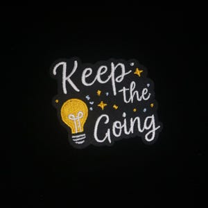 May include: Embroidered patch with the words "Keep the Going" in white script, featuring a yellow lightbulb and small stars. The patch is on a black background.