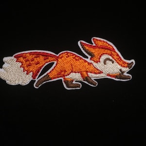 May include: Embroidered patch featuring a cartoon fox in shades of orange, brown, and white. The fox is depicted in a playful pose with a smiling expression. The patch is likely designed for application on clothing or accessories.
