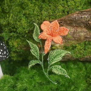 May include: An embroidered orange flower with green leaves and stem. The flower is set against a backdrop of green moss, a wooden log, and a black and white mushroom.