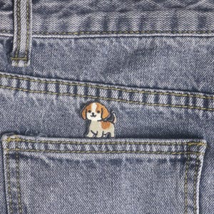 May include: Close-up of blue denim jeans with a small, embroidered puppy patch in the back pocket. The puppy is beige, brown, and white. The jeans have yellow stitching.