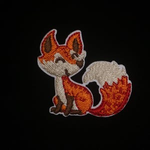 Könnte beinhalten: Gestickter Aufnäher eines Cartoon-Fuchses in Orange-, Braun- und Cremetönen mit weißer Umrandung. Der Fuchs sitzt mit eingerolltem Schwanz. Der Hintergrund ist einfarbig schwarz.
