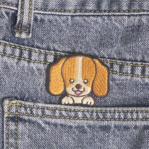 Puede incluir: Parche bordado de un cachorro de dibujos animados con pelaje marrón y blanco, colocado en un bolsillo de mezclilla. El parche presenta una cara de cachorro sonriente con orejas grandes y una lengua rosada. La mezclilla tiene costuras visibles.