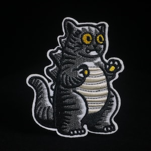 May include: Embroidered patch featuring a cartoon cat-like creature with a Godzilla-inspired design. The patch has a black and white color scheme with yellow accents on the eyes and paws. The creature has a striped belly and a spiky back.