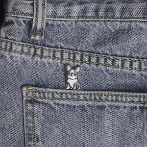 May include: A small, embroidered patch of a French Bulldog dog in black, white, and gray, placed on the pocket of a pair of blue denim jeans. The jeans have yellow stitching and a classic design.