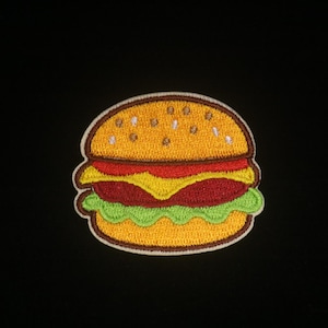 Może przedstawiać: Haftowana naszywka z kreskówkowym hamburgerem. Naszywka posiada żółtą bułkę z ziarnami sezamu, czerwonym pomidorem, żółtym serem i zieloną sałatą. Naszywka ma białą obwódkę i jest umieszczona na czarnym tle.
