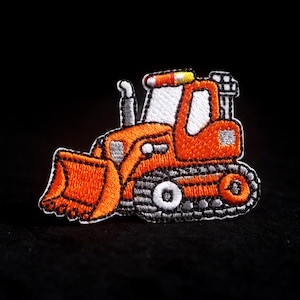 Pode incluir: Patch bordado com uma escavadora de desenho animado laranja, branca e cinzenta. O patch mostra um design detalhado de uma escavadora com uma pá dianteira. Ideal para decorar roupas ou acessórios. O patch está em um fundo preto.