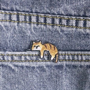 May include: A small, embroidered cat patch in shades of brown and tan, resting on a blue denim pocket. The patch is stitched with black thread, and the denim has yellow stitching.