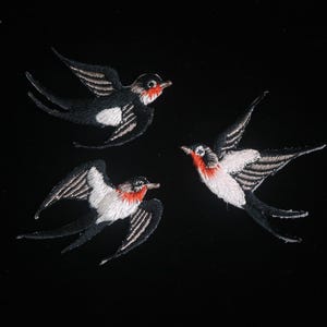 May include: Three embroidered swallows in flight against a black background. The birds feature black and white wings, red throats, and detailed stitching. A decorative element for apparel or home decor.
